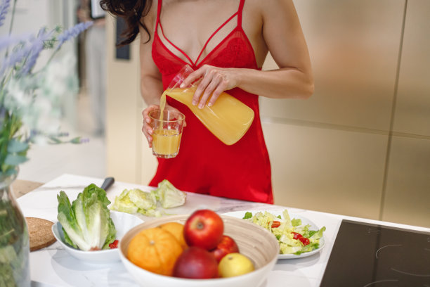 年轻女子在早上拿着玻璃杯倒橙汁的照片。穿着性感的红色睡衣的女人站在厨房里图片下载
