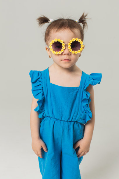 戴着太阳镜和蓝色西装的金发女婴在白色背景的工作室里。的孩子。的童年。发髻式。夏天的时尚图片下载