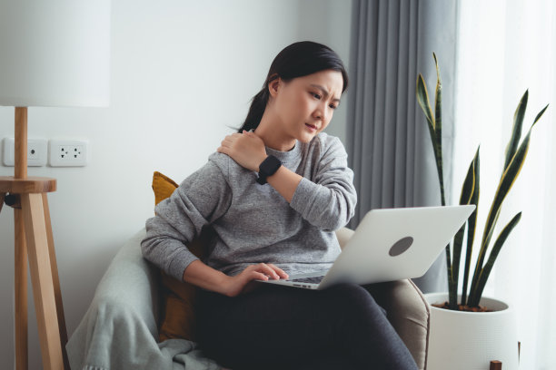 一名亚洲女性坐在家里的客厅里使用笔记本电脑，脖子和肩膀都很痛。图片下载