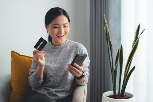 亚洲女性坐在扶手椅上开心地微笑着，用智能手机和信用卡在家里网上购物。图片下载