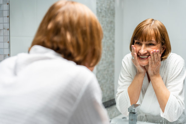 白种老年妇女用面部泡沫和清水洗脸。年长的成熟女性穿着浴袍洗脸，进行健康美容和皮肤护理，然后在浴室里看着镜子图片下载