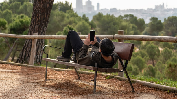 一名男子躺在公园的长椅上休息，一边看手机。图片下载