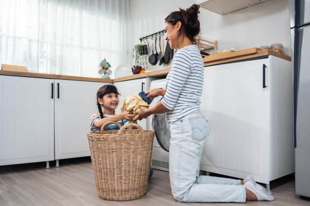 美丽的白人母亲教年幼的女儿洗脏衣服。可爱的小女孩帮助和学习父母妈妈把衣服放在洗衣机电器在家里。Domestic-Housekeeping。图片下载