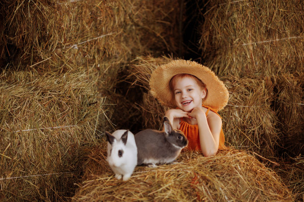 小女孩穿着橙色的裙子，戴着草帽，和一只白色和棕色的兔子坐在稻草捆上图片下载