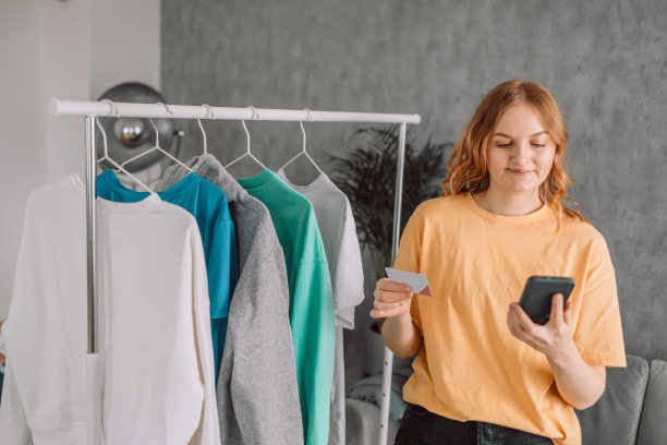 开心的高加索年轻成年妇女使用手机，阅读信息，微笑着站在挂在架子上的衣服旁。图片下载