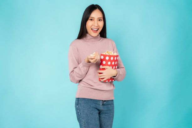 年轻女子拿着一桶红色的爆米花，看着蓝色背景的电影。图片下载