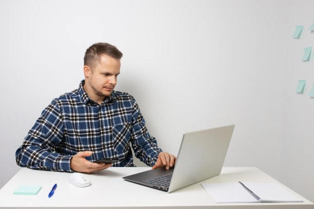 一个年轻人在家庭办公室的画像。使用手机和笔记本电脑。照片侧视图图片下载