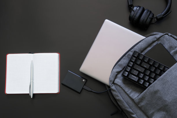 灰色纺织背包或笔记本电脑包随着小工具在一个黑色。银色笔记本电脑，外接ssd，智能手机，耳机，带记事本和游戏键盘的笔。自由职业者和玩家工具。图片下载