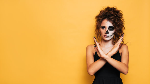 一名年轻女子穿着万圣节服装，在一个黄色的孤立背景上化妆，手里拿着一个头骨。考试或广告的地方。庆祝万圣节的概念。图片下载