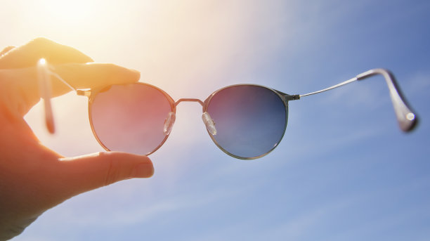 手持时尚的圆形太阳镜对明亮的天空和太阳的背景。在阳光明媚的夏日戴着墨镜。一个男人透过偏光太阳镜看明亮的太阳。夏天的心情图片下载