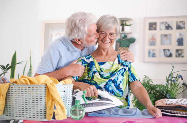 老年妇女微笑着在家里的熨衣板上熨衣服，而她的丈夫温柔地亲吻着这对幸福的退休白人夫妇图片下载