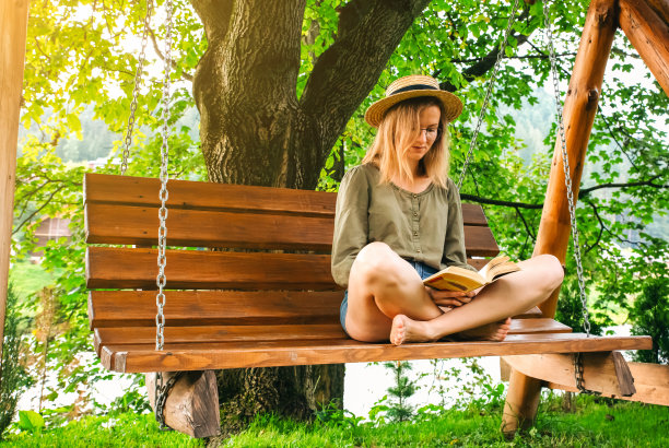 戴眼镜的时髦女孩坐在木凳上看书。女人在户外的门廊上放松，在大自然中荡秋千。舒适的照片。夏天的心情。图片下载