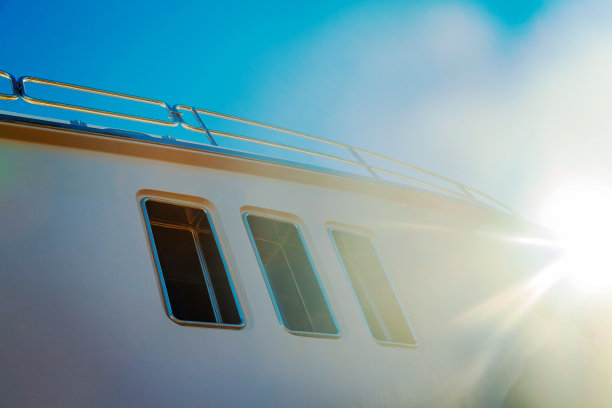 这是一艘豪华的白色游艇的船体，上面有舷窗，映衬着蓝色的天空和角落里的阳光。图片下载