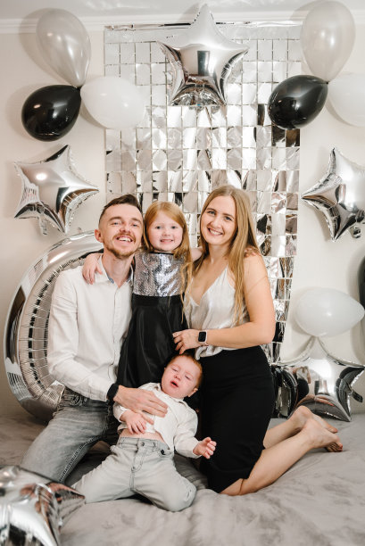 婴儿哭了。幸福的家庭庆祝生日聚会。爸爸妈妈恭贺女儿、儿子生日近拱门。妈妈，爸爸抱着孩子。照片墙装饰白色、黑色、银色气球。图片下载