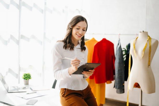 时装设计师女士在现代工作室里用智能手机和笔记本电脑与数码平板电脑交谈，衣服挂在架子上图片下载