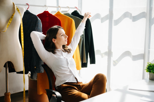 一位年轻的白人女性坐在时装设计师的家庭办公室的桌子上，手里拿着平板电脑、笔记本电脑和智能手机。图片下载