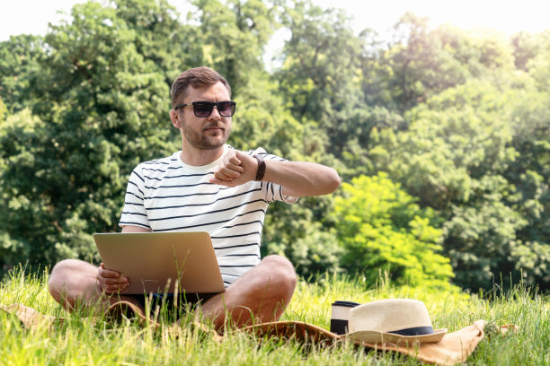 节省时间和计划，男人在笔记本电脑上工作，看着他的手表，而坐在草坪上在公园户外工作。图片下载