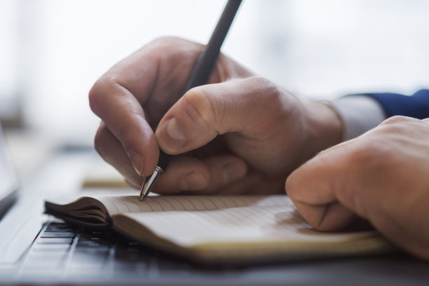 一个男人的手的特写镜头，他在一个光滑的笔记本电脑上的记事本上写字，背景是失焦的图片下载