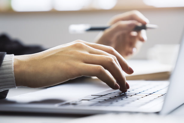 这幅图片描绘了一位女士的手在一台高级笔记本电脑键盘上打字，背景是模糊的办公室环境图片下载