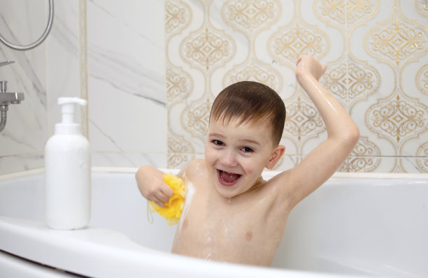 有趣的孩子正在洗澡，坐在有肥皂和洗发水泡沫的白色浴缸里，用他的头发洗他的头。儿童用洗发水、护发产品和肥皂。健康和卫生的概念图片下载