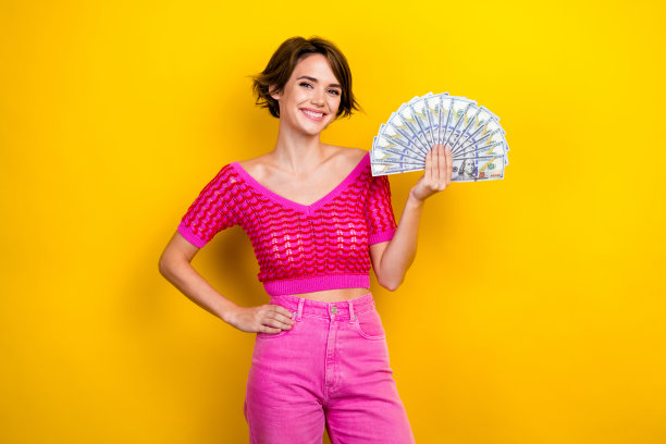 照片微笑的女士鲍勃棕色的头发，穿着粉红色的时尚针织背心牛仔裤自信地拿着她的月收入现金孤立的黄色背景图片下载