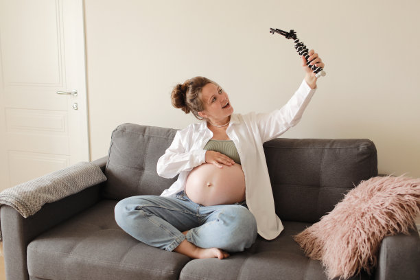 快乐的准妈妈和受欢迎的视频博主坐在现代公寓的沙发上为在线博客录制视频，并对着智能手机摄像头展示她赤裸的大肚子。怀孕与博客概念图片下载