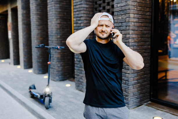 一名成年男子，头戴白色棒球帽，穿着便服，站在电动滑板车旁边，在街上用手机聊天。这个男人心烦意乱，情绪低落图片下载