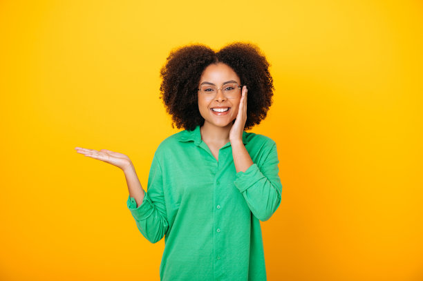 快乐的西班牙裔或巴西人可爱的卷发女人戴着眼镜，穿着绿色衬衫，用手指着旁边的广告空地，对着镜头微笑，站在孤立的橙色背景上图片下载