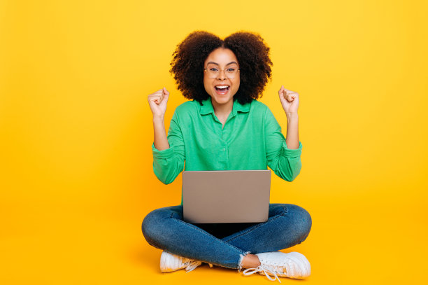 快乐的非裔美国人或巴西卷发女人的全身照片，穿着时髦，坐在黄色背景上，拿着一台笔记本电脑，为成功而高兴，用手比划，对着镜头微笑图片下载