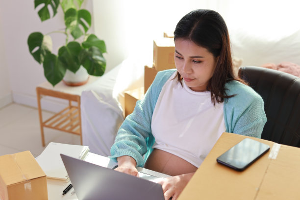 迷人快乐的亚洲孕妇坐着使用移动智能手机，用电脑为客户下单。准妈妈在怀孕期间为婴儿的出生做准备和工作图片下载