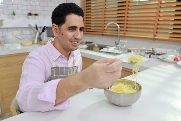 特写年轻快乐的西班牙卷发男人坐在桌子上吃美味的方便面午餐在厨房用筷子和砖墙。不健康和廉价食品的生活方式概念图片下载