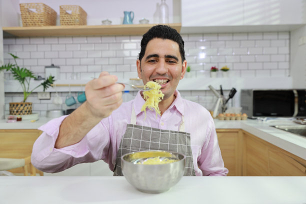 特写年轻快乐的西班牙卷发男人坐在桌子上吃美味的方便面午餐在厨房用筷子和砖墙。不健康和廉价食品的生活方式概念图片下载