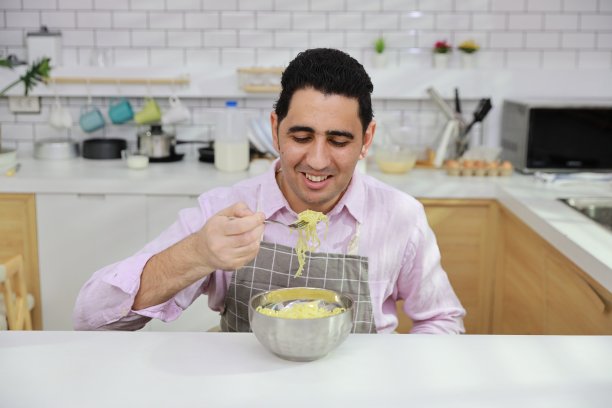 特写年轻快乐的西班牙卷发男人坐在桌子上吃美味的方便面午餐在厨房用筷子和砖墙。不健康和廉价食品的生活方式概念图片下载