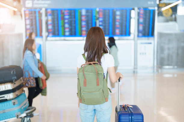 在办理登机手续前，一名年轻女子拿着包和行李在国际机场查看航班时间信息板。旅游，度假，旅行和交通的概念图片下载