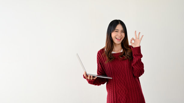 一位微笑着的亚洲女大学生拿着笔记本电脑，做着“ok”的手势图片下载