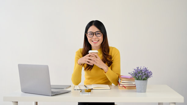 一位快乐的亚洲女职员坐在办公桌前，端着她的咖啡杯。图片下载