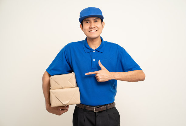 快乐送货亚洲人在蓝色制服站着拿箱子包裹纸板孤立的白色背景。微笑的男服务人员。送货快递及海运服务。图片下载