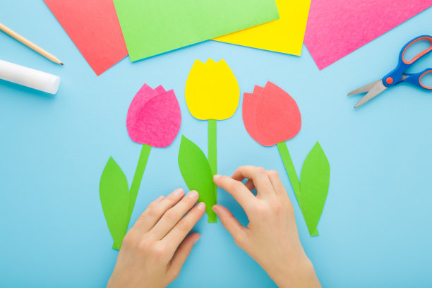年轻的成年妇女双手和五颜六色的郁金香花与绿叶从纸上浅蓝色的桌子背景。柔和的颜色。视角镜头。特写镜头。制作装饰元素。自顶向下视图。图片下载