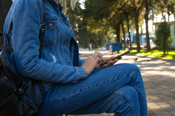 一个女人手持智能手机的特写镜头。一位穿着牛仔裤和牛仔夹克的年轻女子坐在公园的长椅上，一边读着短信，一边用手机看视频。图片下载