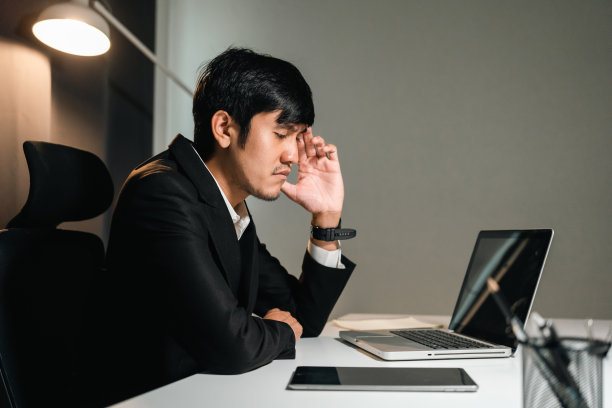 亚洲商人晚上在黑暗的办公室里用笔记本电脑工作。印度人努力加班，认真思考。在深夜温暖的灯光下感到压力和头痛图片下载