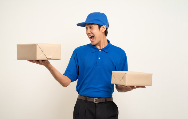 快乐送货亚洲人在蓝色制服站着拿箱子包裹纸板孤立的白色背景。微笑的男服务人员。送货快递及海运服务。图片下载