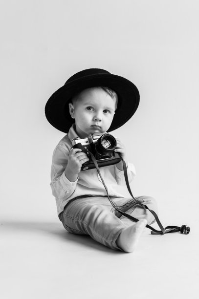 戴帽子的孩子坐在地板上玩照相机。世界摄影师日。有趣的孩子拿着复古的老式相机孤立在白墙上。孩子们的工作室肖像。模型。黑白照片。图片下载