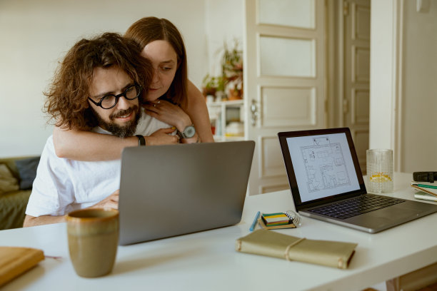 年轻的微笑男子在笔记本电脑上工作，女友在家里拥抱他。远程工作概念图片下载