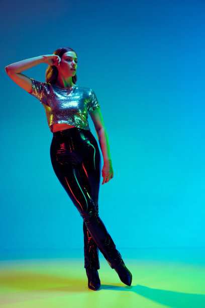 高级时装模特，年轻女子穿着时髦的衣服，黑色皮裤和银色上衣，在霓虹灯的蓝色背景下摆姿势。概念之美，时装周图片下载