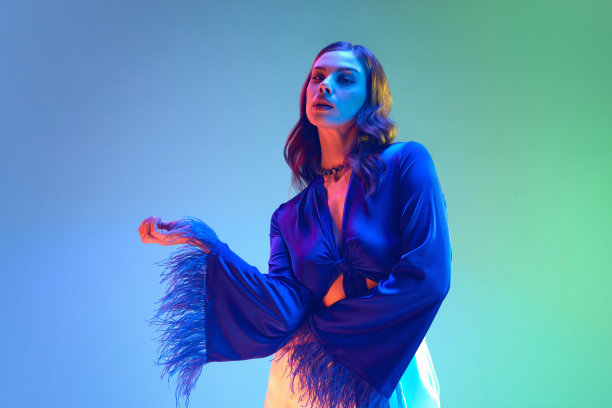 漂亮性感的女人，深色头发，波浪状闪亮的头发，穿着蓝色丝绸衬衫，在渐变的霓虹背景上有羽毛。魅力舒适家装，奢华睡衣图片下载