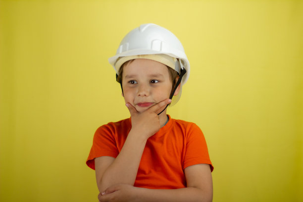 可爱的孩子认为他的下巴在一个建筑头盔。孤立在黄色背景上。一个戴着头盔的小工程师轻轻地揉着下巴。儿童从事成人职业的概念。图片下载