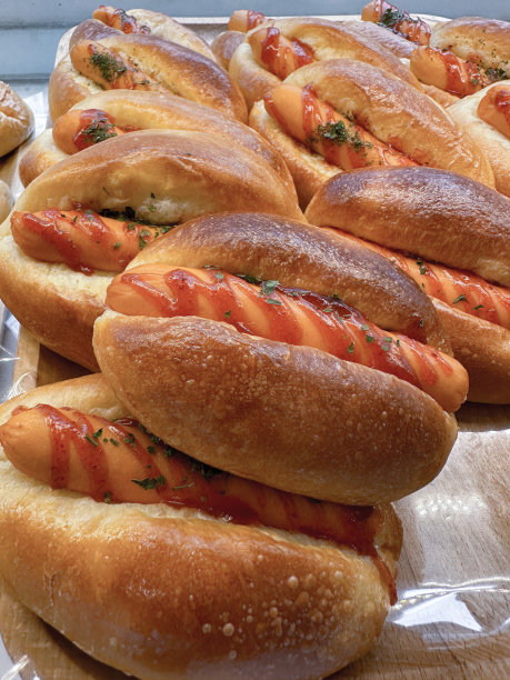 这批面包手指卷的特写图片，里面装满了热狗香肠，上面覆盖着番茄酱，堆放在面包店展示的木架子上，重点放在前景上图片下载