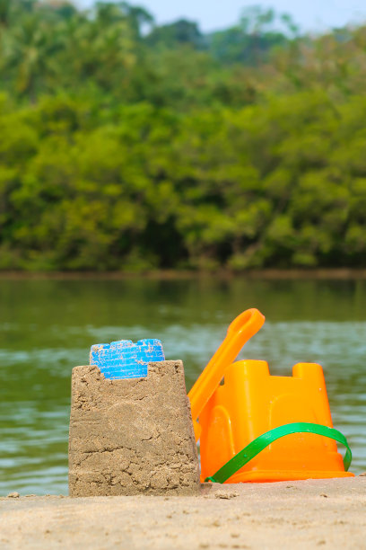 特写照片中，一个无法辨认的人穿着冲浪短裤和条纹短袖衬衫，站在泻湖沙滩上齐脚踝深的水里，用塑料堆沙堡，色彩鲜艳的儿童海滩玩具，水桶和铲子，林地景色图片下载