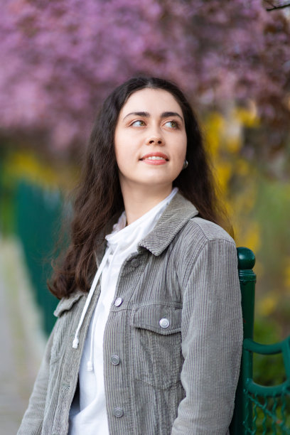 年轻美丽的肖像，微笑的白人妇女穿着夹克在公园摆姿势。盛开的树在背景。垂直的。无忧无虑的心理概念图片下载