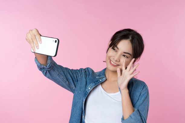 年轻的亚洲女子在粉红色的背景下用智能手机自拍。图片下载